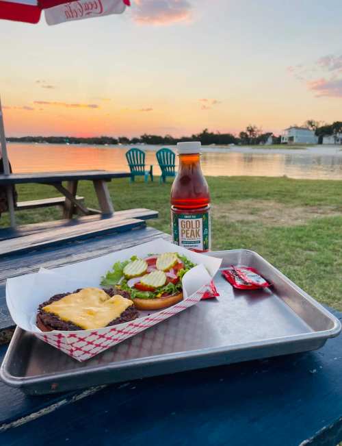 A burger with cheese, lettuce, and tomato on a tray, alongside a bottle of iced tea, with a sunset over a lake in the background.
