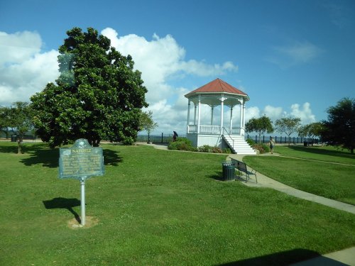 A white gazebo on a grassy area with trees, a historical marker, and a view of the water under a blue sky.