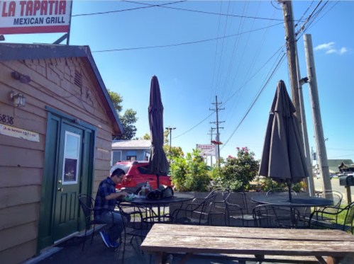 A man sits at an outdoor table near a Mexican grill, with umbrellas and a clear blue sky in the background.
