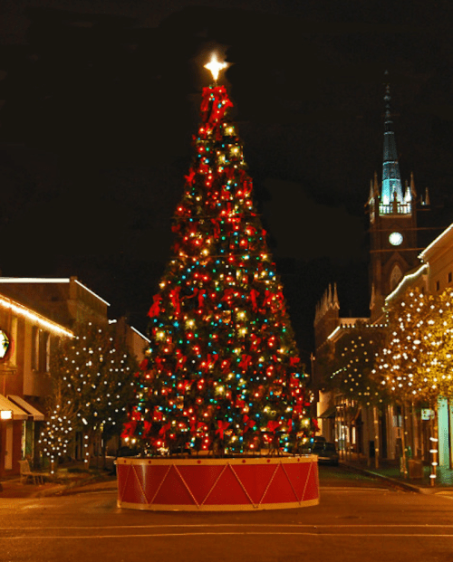A brightly lit Christmas tree adorned with red bows and colorful lights, set against a night backdrop of a town square.