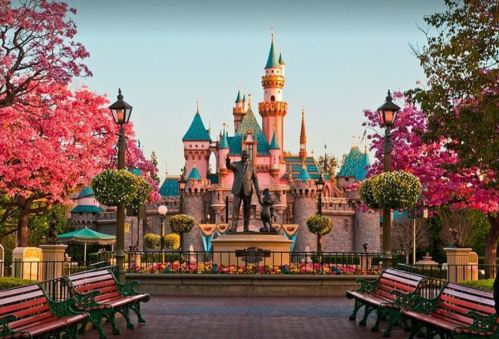 A colorful castle surrounded by blooming trees and benches, with a statue in the foreground at a theme park.
