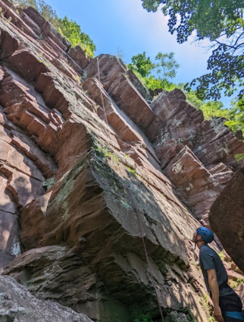 A climber gazes up at a steep rock face, preparing to ascend under a clear blue sky and lush greenery.