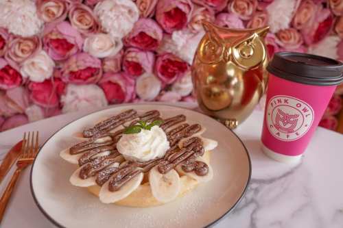 A plate of banana pancakes topped with chocolate drizzle and whipped cream, next to a pink coffee cup and a gold owl decoration.
