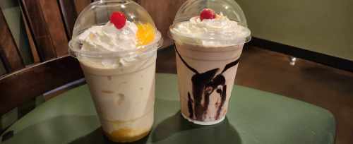 Two milkshake cups topped with whipped cream and cherries, one with fruit and the other with chocolate drizzle, on a table.