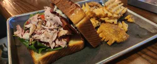 A hearty sandwich with layers of meat and greens, served with waffle fries on a metal tray.