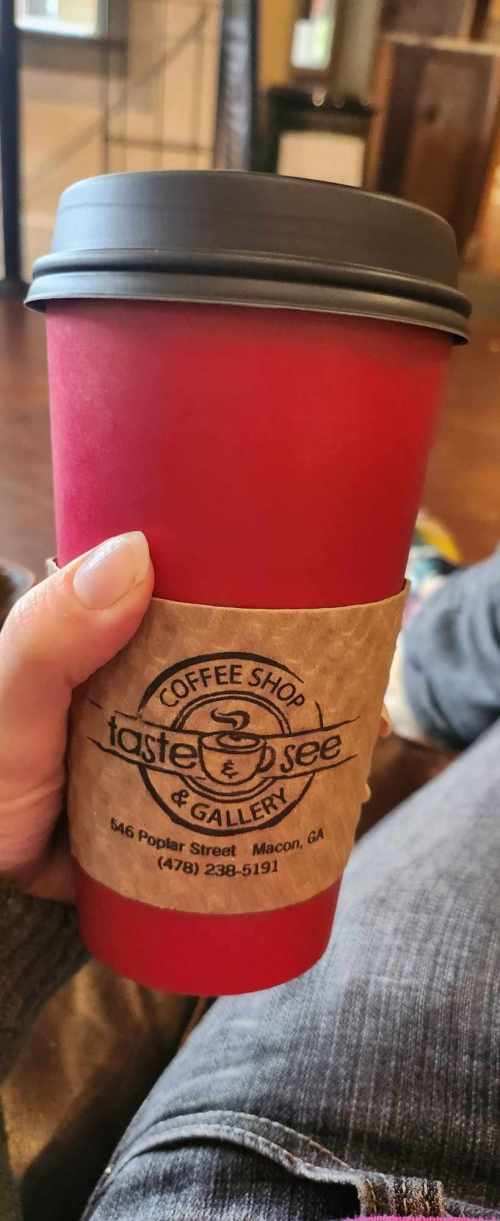 A hand holding a red coffee cup with a brown sleeve, featuring the logo of a coffee shop and gallery.