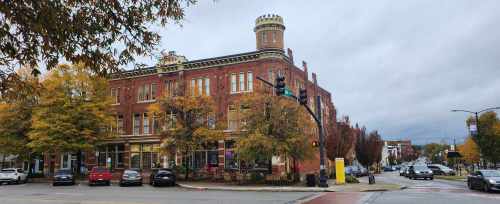Historic brick building with a turret, surrounded by autumn trees and parked cars on a cloudy day.