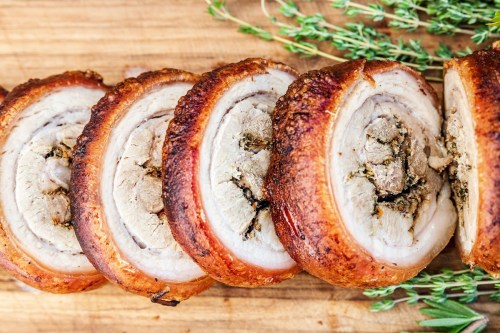Sliced roasted meat with crispy skin, garnished with fresh thyme on a wooden cutting board.