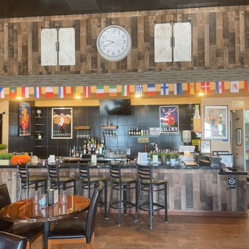 A cozy bar area with a wooden decor, clock, flags, and framed posters, featuring a well-stocked counter and seating.