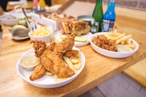 Two plates of crispy fried chicken tenders with crinkle-cut fries and dipping sauce, alongside colorful drinks.