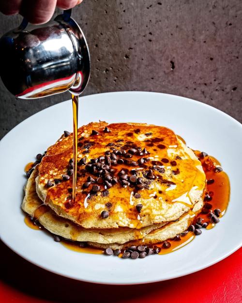 A stack of pancakes topped with chocolate chips, drizzled with syrup, on a white plate against a textured background.