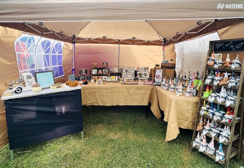 A craft market stall featuring handmade items, including decorations and gifts, under a tent with a grassy floor.