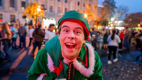A cheerful person in an elf costume smiles widely at a festive outdoor event with a crowd in the background.