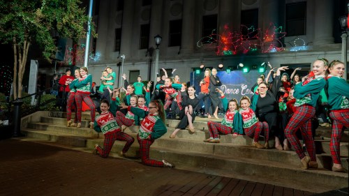 A group of dancers in festive outfits poses on steps, with holiday lights and decorations in the background.