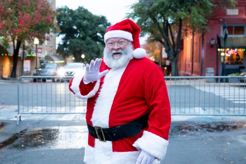 A cheerful Santa Claus in a red suit waves, standing in a festive outdoor setting with holiday decorations.