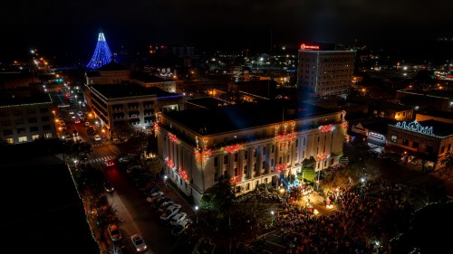 Aerial view of a festive city at night, featuring a decorated building, a large Christmas tree, and a crowd gathered below.