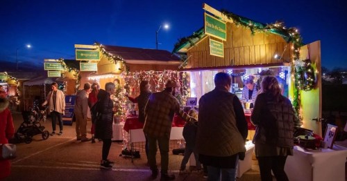 A festive market scene at night with decorated stalls, people shopping, and holiday lights illuminating the area.