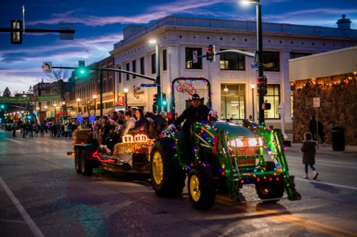A festive tractor with lights pulls a decorated float through a city street during a parade at dusk.