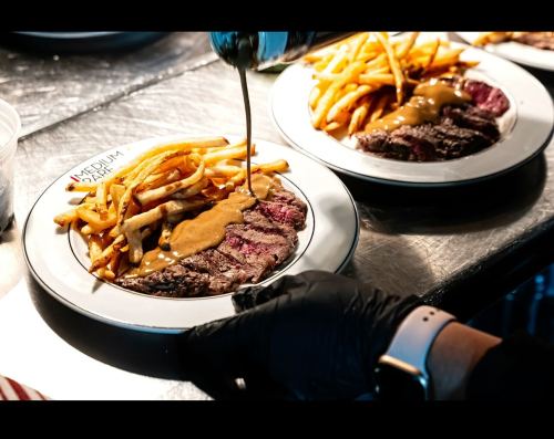 A plate of medium steak with gravy and crispy fries, being drizzled with sauce by a gloved hand.
