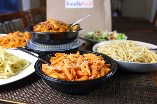 A variety of pasta dishes in black and white bowls, with a salad and a takeout bag in the background.
