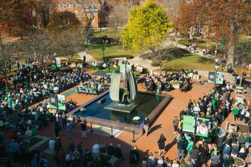 A large crowd gathers around a fountain in a park, surrounded by trees and buildings, for an outdoor event.