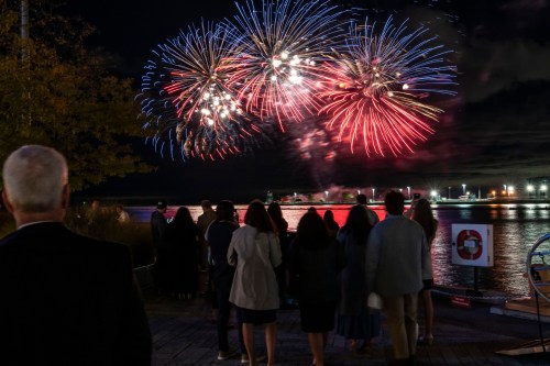 A group of people watches colorful fireworks explode over a waterfront at night.