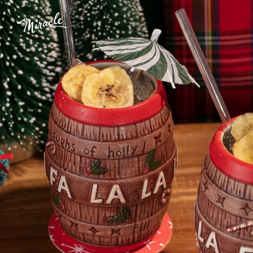 Two festive cocktails in decorative barrels, topped with banana slices and a striped umbrella, set against a holiday backdrop.