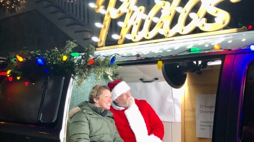A man in a Santa suit and a woman smile together in front of a festive photo booth with lights.