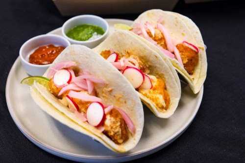 Three fish tacos on a plate, topped with pickled onions and radishes, served with salsa and green sauce on the side.