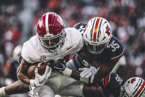 A football player in a red Alabama jersey is tackled by an opponent in a gray Auburn jersey during a game.