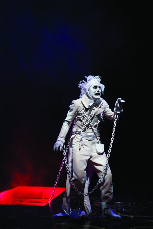 A ghostly figure in chains stands dramatically on stage, illuminated by red light against a dark background.