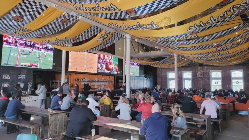 A lively indoor venue with large screens showing sports, decorated with Oktoberfest banners and filled with patrons.