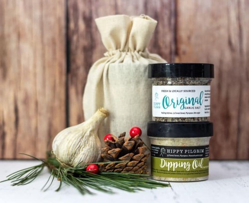 A burlap bag, jars of garlic salt and dipping oil, a garlic bulb, pinecones, and pine branches on a wooden background.
