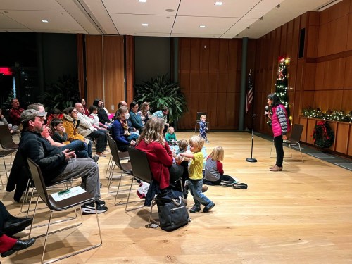 A speaker addresses an audience in a festive room, with children sitting on the floor and decorations in the background.