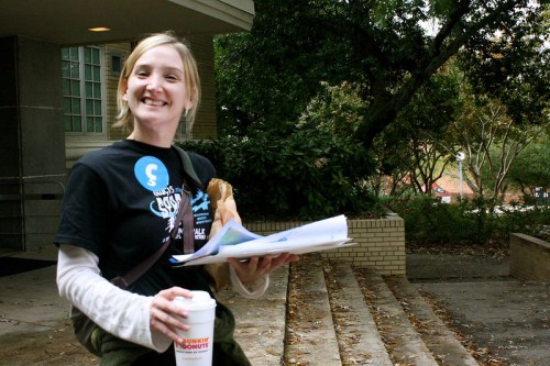 Smiling person holding papers and a coffee cup, standing outside near steps and greenery.