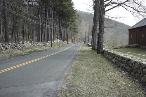 A quiet, winding road lined with trees and stone walls, leading into a rural landscape with a distant building.