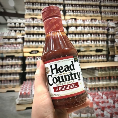 A person holding a bottle of Head Country Original Bar-B-Q Sauce in a warehouse filled with stacked products.