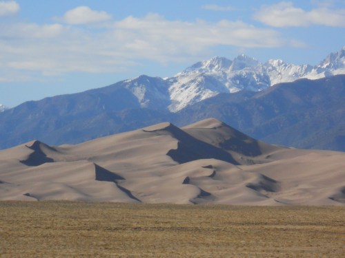 A vast desert landscape with rolling sand dunes in the foreground and snow-capped mountains in the background under a blue sky.