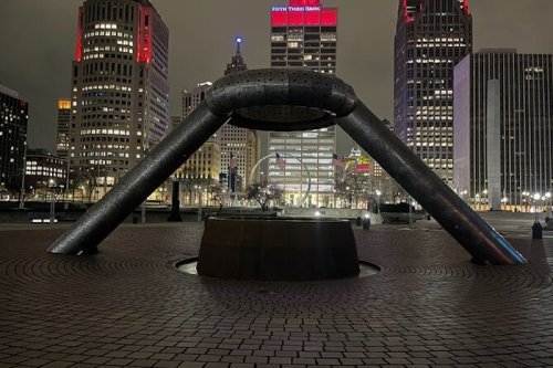 A modern sculpture with a curved design in a city plaza, surrounded by tall buildings and illuminated at night.