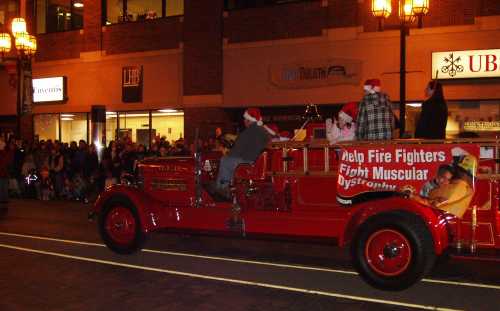 A vintage fire truck in a parade, decorated for the holidays, with spectators watching from the sidewalk.