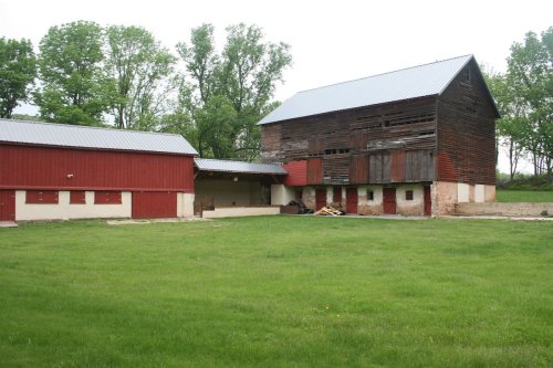 A rustic farm scene featuring a large, weathered barn and a smaller red building, surrounded by green grass and trees.
