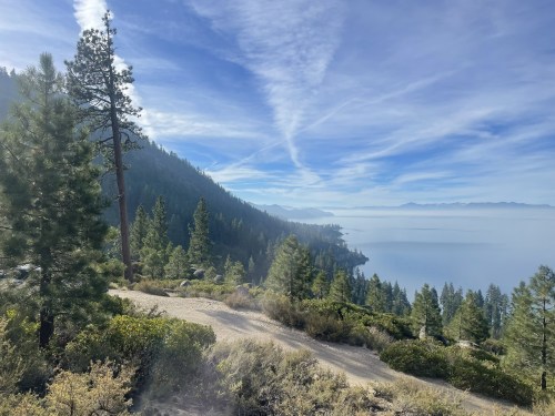 A scenic view of a misty lake surrounded by mountains and pine trees under a blue sky with wispy clouds.