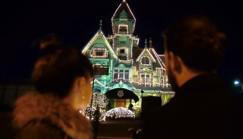 A couple admires a beautifully lit Victorian-style house decorated with holiday lights at night.