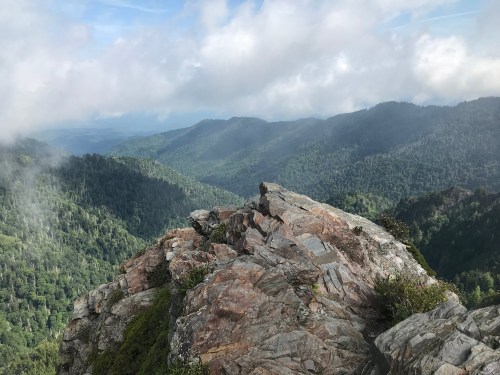A rocky outcrop overlooks a lush green mountain range under a cloudy sky.