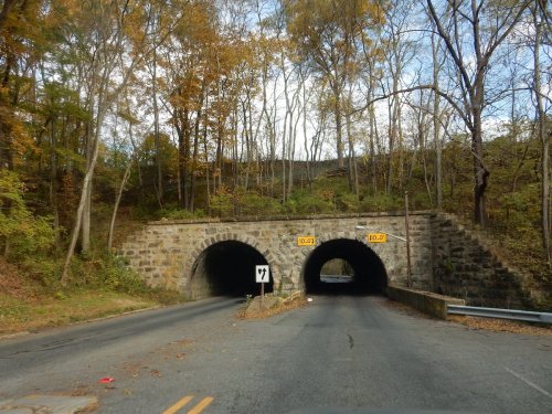 Two stone arch tunnels under a tree-lined road, surrounded by autumn foliage. Road signs indicate tunnel heights.