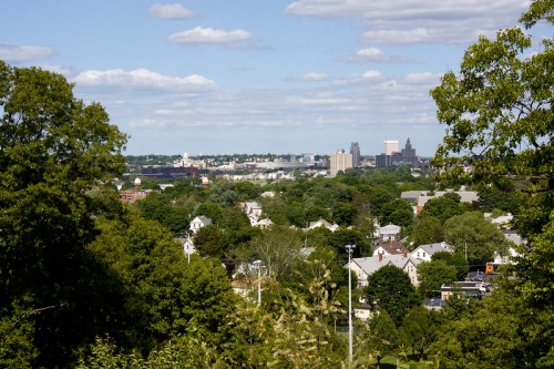 A panoramic view of a city skyline surrounded by lush greenery and scattered residential buildings under a blue sky.
