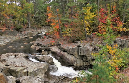 A serene river flows through rocky terrain, surrounded by vibrant autumn foliage in shades of red, orange, and yellow.