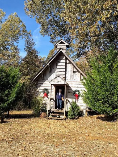 A small wooden chapel with a peaked roof, surrounded by trees and autumn leaves, with a person standing at the entrance.