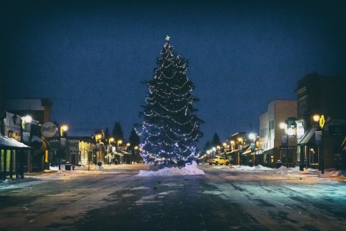 A snowy street at night featuring a large, decorated Christmas tree and illuminated storefronts.