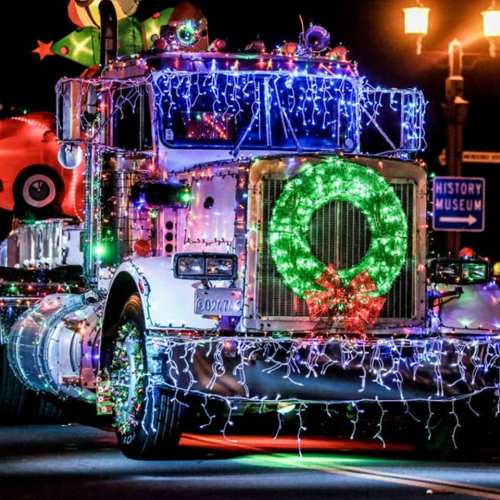 A festive truck adorned with colorful lights and a wreath, parked near a museum during a holiday celebration.